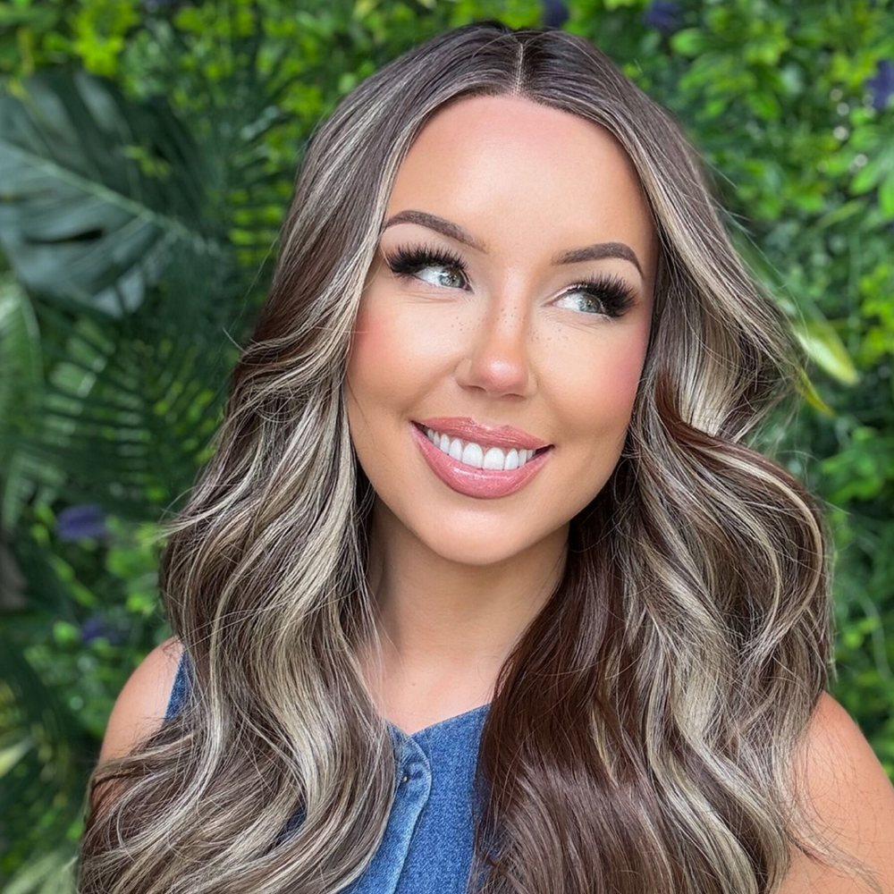 A smiling woman wearing a long, layered brunette wig with blonde highlights, photographed against a wall of green leaves.